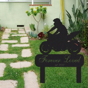 Puede incluir: Silueta negra de metal de una persona conduciendo una motocicleta con las palabras "Forever Loved est. 2023" en un letrero debajo. La silueta está frente a un césped verde y un camino de piedra que conduce a una casa.
