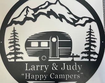 Könnte beinhalten: Schwarze Metallsilhouette eines Wohnwagens vor Bergen mit zwei Kiefern auf jeder Seite. Der Text "Larry & Judy" und "Happy Campers" befindet sich unter dem Wohnwagen.
