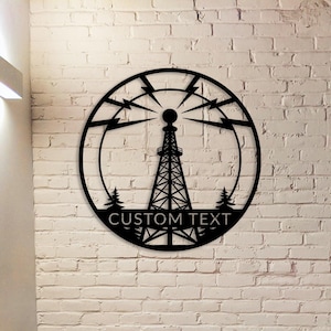 Pode incluir: Uma peça de arte de parede em metal preto com um design de torre de rádio dentro de uma moldura circular. O design inclui raios, árvores e o texto "CUSTOM TEXT" na parte inferior. A obra de arte é montada em uma parede de tijolos brancos.