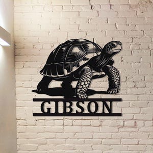 May include: Black metal turtle wall art with the name "GIBSON" below. The turtle has a detailed shell and textured legs. The art is mounted on a white brick wall.