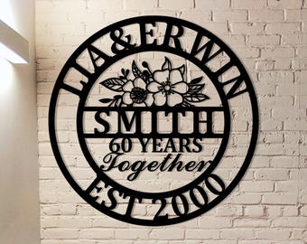 Könnte beinhalten: Schwarze Metall-Wandkunst mit den Namen "LIA & ERWIN" in einem kreisförmigen Design. Das Kunstwerk enthält den Familiennamen "SMITH", den Text "60 YEARS Together" und das Jahr "EST 2000". Florale Akzente sind ebenfalls enthalten.