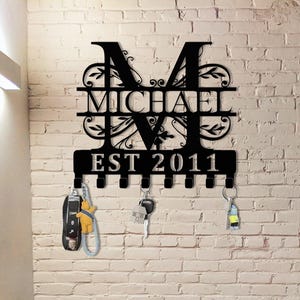 May include: Black metal key holder with the name "MICHAEL" and the year "EST 2011" cut out. The key holder has an ornate design with the letter "M" at the top and several hooks for keys. The key holder is mounted on a white brick wall.