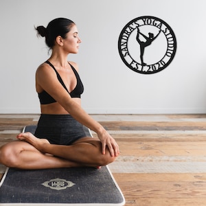 Może przedstawiać: Kobieta siedzi w pozycji jogi na czarnej macie do jogi z białym obramowaniem. Mata ma ozdobny wz&oacute;r w środku. Czarny metalowy znak z tekstem "Sandra's Yoga Studio Est. 2020" i sylwetką kobiety w pozycji jogi znajduje się na ścianie za nią.