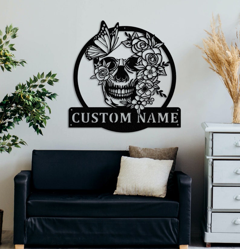 May include: Black metal wall decor featuring a skull with floral accents and a butterfly. The skull has a floral design covering the eyes and a butterfly perched on the top of the skull. The text "CUSTOM NAME" is displayed on a separate metal plaque below the skull.