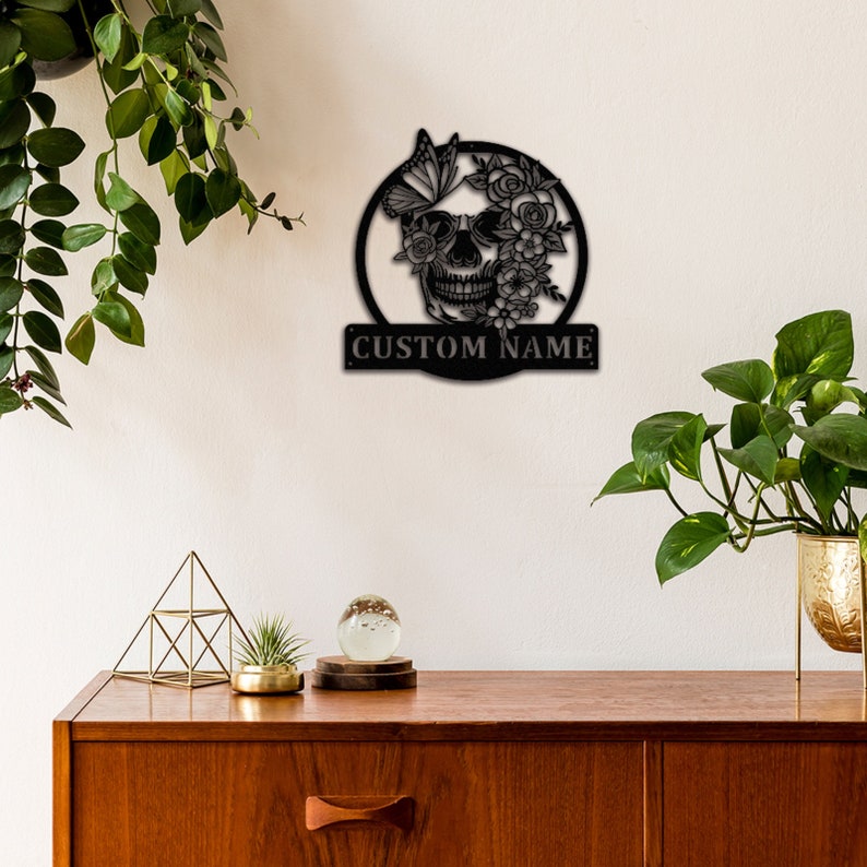 May include: Black metal wall decor featuring a skull with flowers and a butterfly. The skull is surrounded by a circle and the text "CUSTOM NAME" is below the circle.
