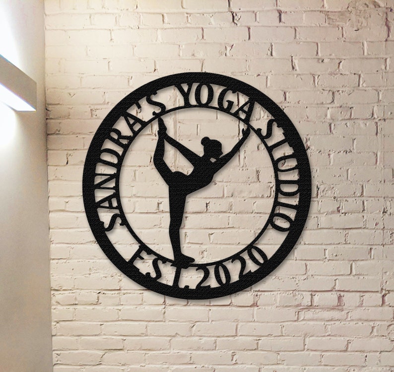 Może przedstawiać: Czarny metalowy znak z sylwetką pozycji jogi w środku. Znak głosi "Sandra's Yoga Studio Est. 2020".