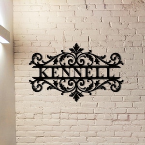 Può includere: Arte da parete in metallo nero con il nome "KENNELL" in un carattere decorativo. Il nome è centrato all'interno di un ornato disegno a volute. L'arte è montata su un muro di mattoni bianchi.