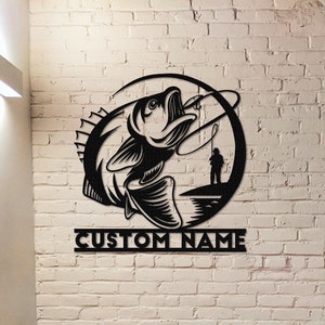 May include: Black metal wall art featuring a silhouette of a fish jumping out of the water with a fishing line and a fisherman in the background. The art is in a circle shape with the text "CUSTOM NAME" below.