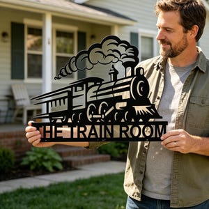 May include: A black metal wall sign featuring a silhouette of a steam locomotive with smoke billowing from the chimney. The text THE TRAIN ROOM is cut out in bold capital letters along the bottom edge of the rectangular metal piece.