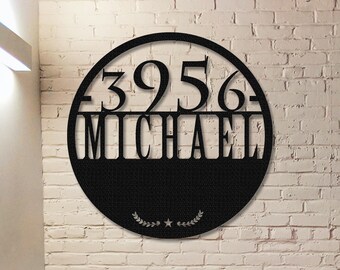 Puede incluir: Letrero de dirección de metal negro con el número "3956" sobre el nombre "MICHAEL" dentro de un marco circular. El letrero está montado en una pared de ladrillos blancos. Una corona de laurel decorativa y una estrella están en la parte inferior.