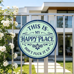 May include: A round sign with the text "THIS IS MY HAPPY PLACE DON'T FUCK IT UP" in a dark blue font on a light green background. The sign is mounted on a silver metal fence, with white flowers in the background.