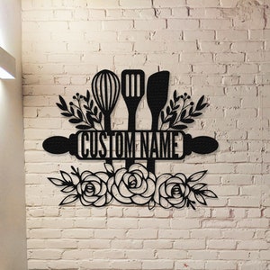Peut inclure: Décoration murale de cuisine en métal noir avec un rouleau à pâtisserie, un fouet, une spatule et des accents floraux. Le rouleau à pâtisserie porte le texte "CUSTOM NAME".