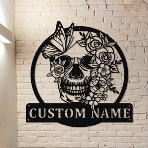 May include: Black metal wall art featuring a skull with flowers and a butterfly. The skull is surrounded by a circle and the text "CUSTOM NAME" is below the circle.