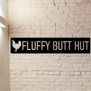 Op de afbeelding: Een zwart metalen bord met de witte tekst "FLUFFY BUTT HUT" en een silhouet van een kip. Het bord is bevestigd aan een witte bakstenen muur, wat een rustieke en humoristische uitstraling creëert.