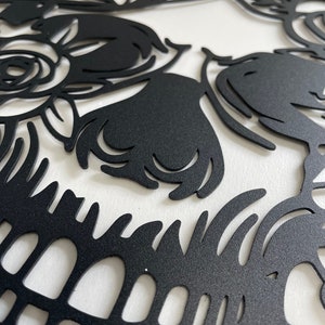 May include: Black and white sugar skull design with intricate floral details. The skull is cut out of a black material and mounted on a white background.