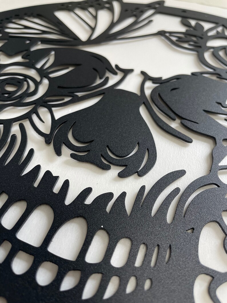 May include: Black and white sugar skull design with intricate floral details. The skull is cut out of a black material and mounted on a white background.