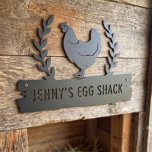 May include: A matte black metal sign featuring a cutout of a chicken flanked by two leafy branches. The sign has a rectangular base with a rough, wood-like edge and the text JENNY'S EGG SHACK cut out in the center, secured by two visible screws.