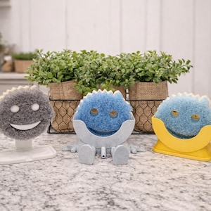 May include: Three whimsical kitchen sponges in the shape of smiling faces. One is gray with a white base, the second is blue with a gray holder, and the third is blue with a yellow holder. Green plants in burlap pots are in the background.