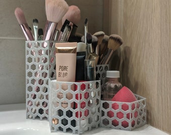 May include: A set of three gray hexagonal makeup organizers with a variety of makeup brushes, a pink makeup sponge, and a clear bottle with a pink liquid. The organizers are filled with makeup brushes, a pink makeup sponge, and a clear bottle with a pink liquid. The organizers are made of a gray, hexagonal material. The text "PORE BLUR" is visible on a pink tube of makeup.
