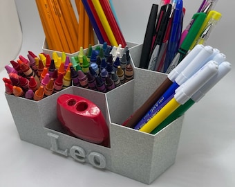 May include: A gray hexagonal pencil holder with six compartments. The holder is filled with various art supplies, including crayons, colored pencils, markers, and pens. The holder has the name "Leo" printed on the front.