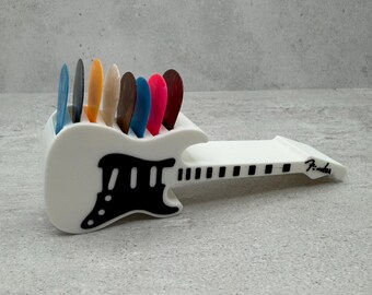 May include: A white guitar pick holder with a black pickguard design, holding a variety of colorful guitar picks. The holder is shaped like a guitar and has the word "Fender" printed on the side.