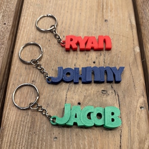 May include: Three personalized keychains with names: "RYAN" in red, "JOHNNY" in blue, and "JACOB" in green. Each keychain has a silver-colored ring and chain. The keychains are displayed on a wooden surface.