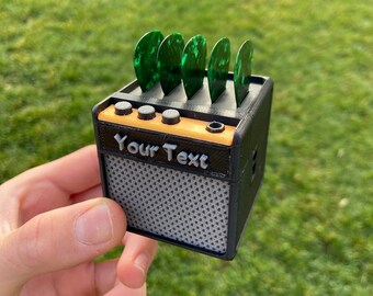 May include: A small black and orange 3D printed guitar amplifier with a gray speaker grill and a slot for guitar picks. The amplifier has the text "Your Text" printed on the front.