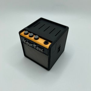 May include: A small black and gray guitar amplifier with a wood panel and four knobs. The amplifier has a speaker grill and the text "Your Text" printed on the front.