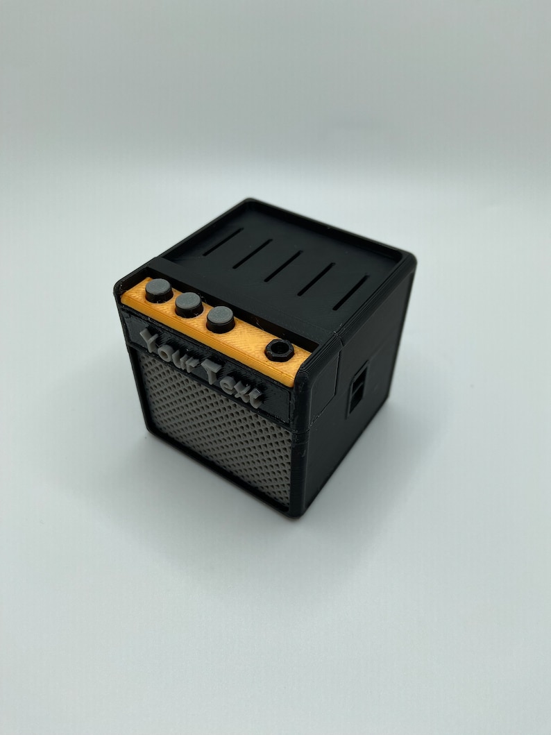 May include: A small black and gray guitar amplifier with a wood panel and four knobs. The amplifier has a speaker grill and the text "Your Text" printed on the front.