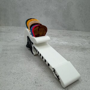 May include: A white guitar pick holder with a black and white design, holding a variety of colorful guitar picks. The holder has the word "Fender" printed on it.