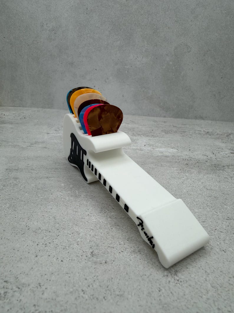 May include: A white guitar pick holder with a black and white design, holding a variety of colorful guitar picks. The holder has the word "Fender" printed on it.