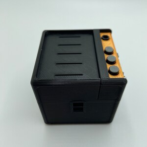 May include: A black and brown miniature stove with four black knobs and a small window. The stove is made of plastic and has a textured surface.