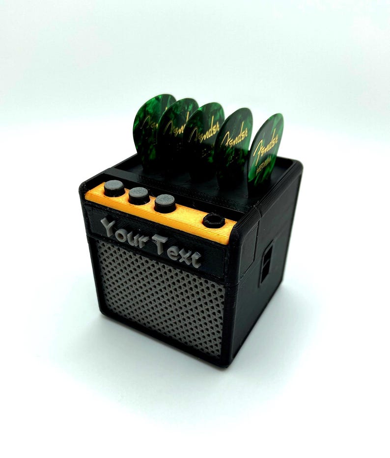 May include: A black and yellow miniature guitar amplifier with a speaker grill and four green guitar picks on top. The amplifier has a label that says "Your Text".