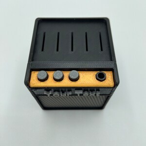 May include: A black and gold miniature guitar amplifier with four knobs and a speaker grill. The amplifier has the text "Your Text" printed on the front.