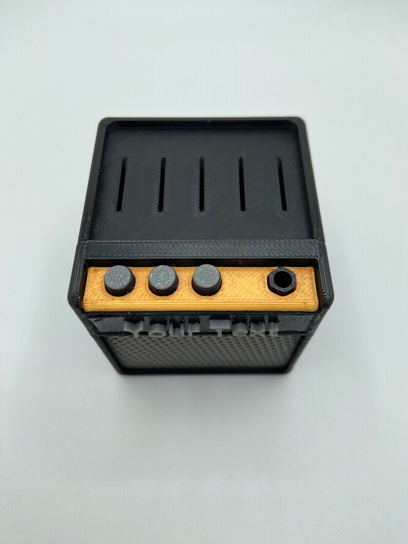May include: A black and gold miniature guitar amplifier with four knobs and a speaker grill. The amplifier has the text "Your Text" printed on the front.