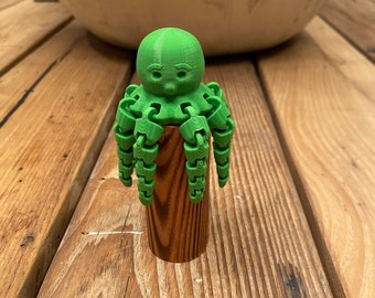 May include: A green octopus figurine with a face and eight articulated arms, standing on a brown wooden cylinder.