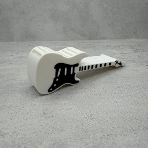 May include: A white guitar-shaped object with black accents, including the pickguard and fretboard. The word "Fender" is printed on the headstock. The object is resting on a gray surface.