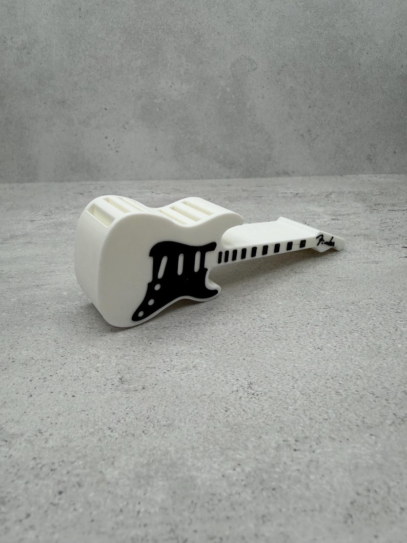 May include: A white guitar-shaped object with black accents, including the pickguard and fretboard. The word "Fender" is printed on the headstock. The object is resting on a gray surface.