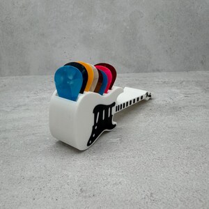 May include: A white guitar pick holder with a black guitar-shaped cutout, holding several colorful guitar picks. The picks are blue, black, orange, brown, pink, and red. The holder has the word "Fender" printed on it.