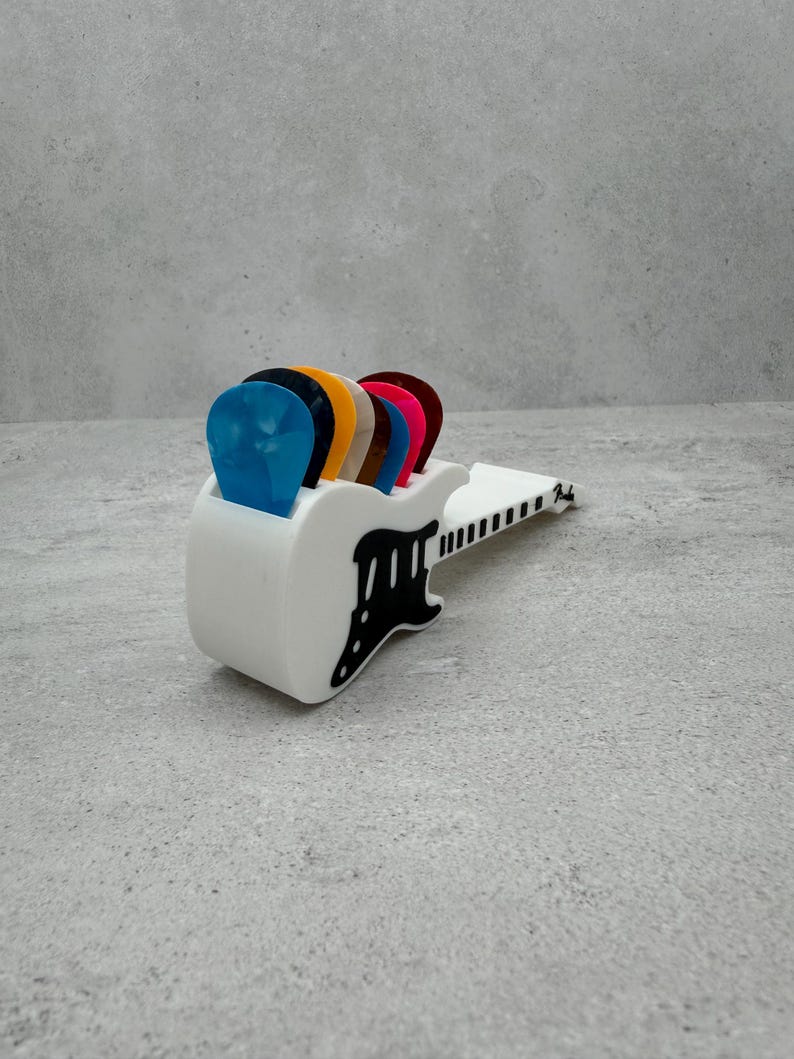 May include: A white guitar pick holder with a black guitar-shaped cutout, holding several colorful guitar picks. The picks are blue, black, orange, brown, pink, and red. The holder has the word "Fender" printed on it.