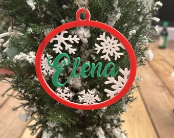 May include: A red and white Christmas ornament with the name "Elena" in green script. The ornament is round with a snowflake design and is hanging from a twine string. The ornament is displayed on a small Christmas tree.