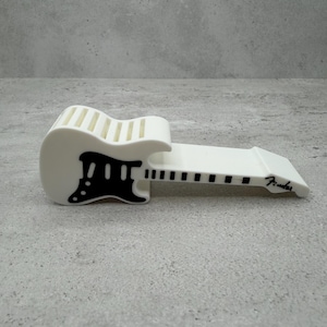 May include: A white guitar-shaped phone stand with black accents. The stand features a detailed design of a guitar, including the body, neck, and headstock. The word "Fender" is printed on the stand.