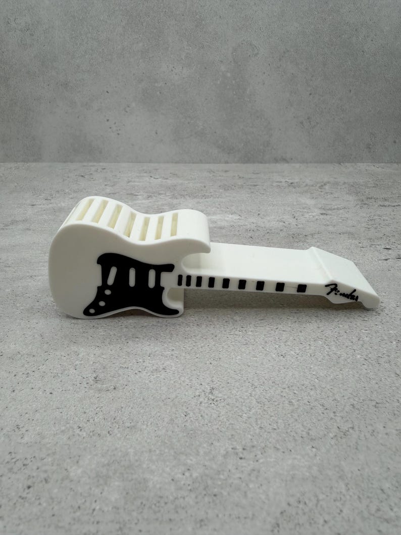 May include: A white guitar-shaped phone stand with black accents. The stand features a detailed design of a guitar, including the body, neck, and headstock. The word "Fender" is printed on the stand.