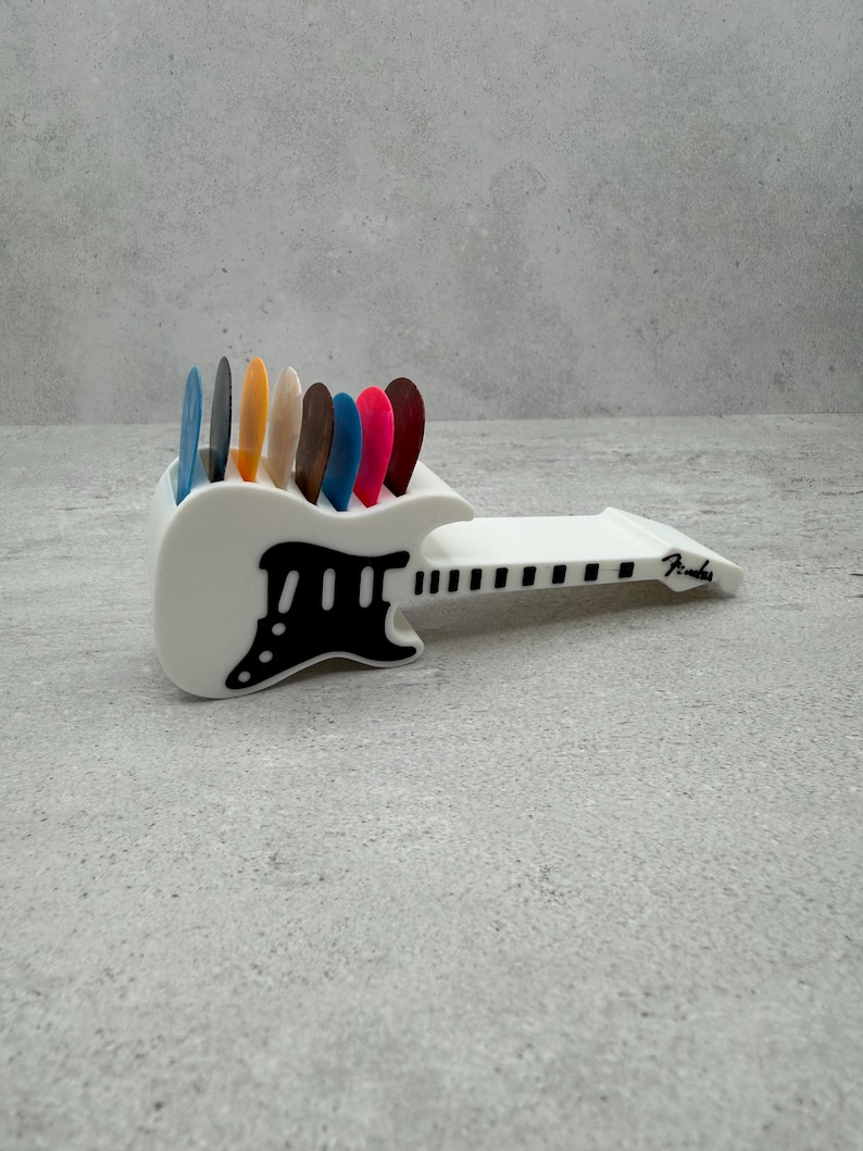 May include: A white guitar pick holder with a black pickguard design, holding a variety of colorful guitar picks. The holder is shaped like a guitar and has the word "Fender" printed on the side.