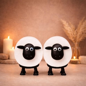 Op de afbeelding: Twee decoratieve schapenbeeldjes. Elk schaap heeft een witte, cirkelvormige body met een zwart gezicht, oren en poten. De schapen hebben grote, ronde ogen. De beeldjes staan tegen een warme, neutrale achtergrond met kaarsen en gedroogde planten.