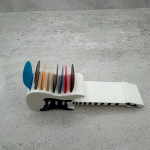 May include: A white guitar pick holder with a black design and the word "Fender" printed on the side. The holder is filled with colorful guitar picks in various shapes and colors, including blue, orange, and red.
