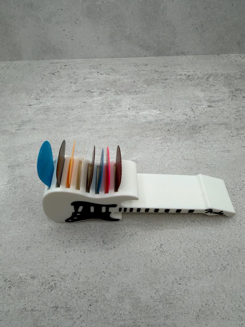 May include: A white guitar pick holder with a black design and the word "Fender" printed on the side. The holder is filled with colorful guitar picks in various shapes and colors, including blue, orange, and red.