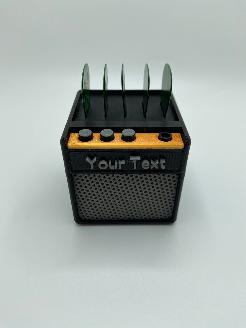 May include: Black and gold guitar pick holder shaped like a miniature amplifier. The holder has six slots for guitar picks and the text "Your Text" is printed on the front.