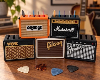 May include: Five rectangular guitar pick holders designed to resemble miniature amplifiers. The holders feature the brand names Orange, Marshall, Vox, Gibson, and Fender. Each unit has slots on top for holding guitar picks, with several colorful picks inserted and four loose picks resting in front.