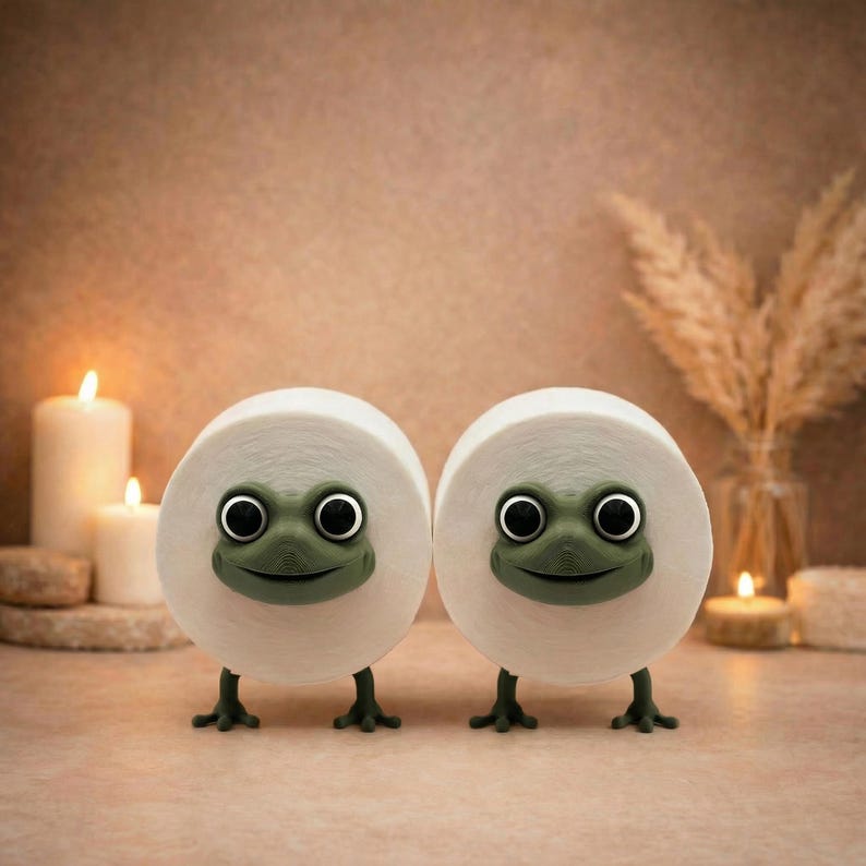 May include: Two whimsical toilet paper roll holders, each designed as a frog with large black eyes and a green face. The rolls are white, and the frog legs are green. The background includes candles and decorative elements, creating a playful bathroom decor theme.